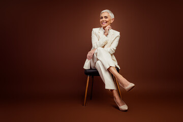 Successful businesswoman in stylish formal attire seated gracefully against a brown background,...