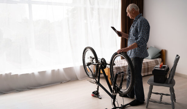 An elderly man repairs a bicycle in an apartment. A passionate Caucasian pensioner does maintenance on his own, stands near the window and holds a tablet in his hands. - Powered by Adobe