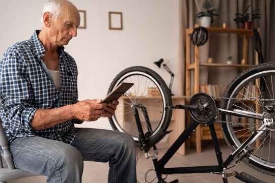 An elderly Caucasian pensioner is enthusiastically repairing his bicycle at home. The man is looking at the tablet intently while sitting near the bicycle, the concept of studying video lessons online - Powered by Adobe