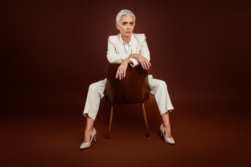 Professional mature businesswoman posing confidently in an elegant suit against a solid brown...