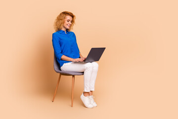 Young female professional seated on a beige background working on a laptop, wearing a blue shirt and white trousers