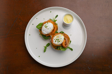 Two poached eggs for brunch on a bed of fresh greens, garnished with herbs, served with a side of creamy sauce on a white plate.