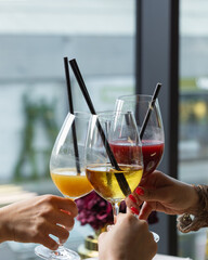 Three hands raise elegant glasses filled with vibrant drinks. The gathering occurs indoors,...