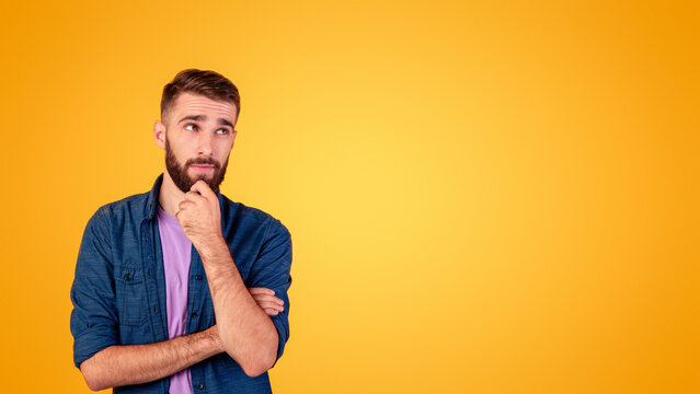 Fototapeta Portrait of attractive young guy thinking and touching his chin over orange studio background, copy space. Pensive millennial man having doubts, making decision or choice, dreaming about something