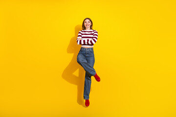 Confident young woman posing against yellow background wearing trendy striped pullover and jeans with a cheerful expression.
