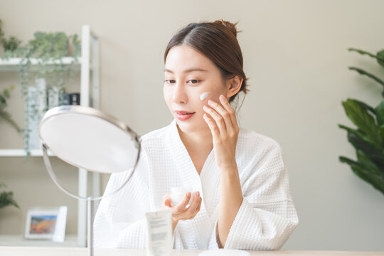 Facial beauty skin care, smile of pretty asian young woman in bathrobe looking at mirror, hand applying moisturizer lotion on her face, putting cream treatment before makeup cosmetic routine at home. - Powered by Adobe