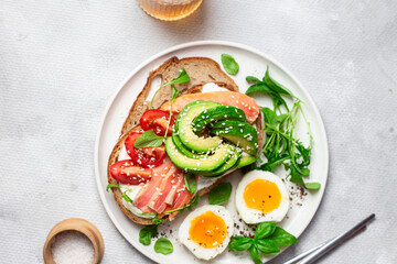  Sandwich with salmon, avocado, tomatoes and egg in a white plate on the table close-up. Healthy breakfast