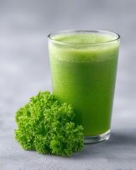 A glass of fresh green juice with frothy top sits next to a bunch of curly parsley on a grey surface.