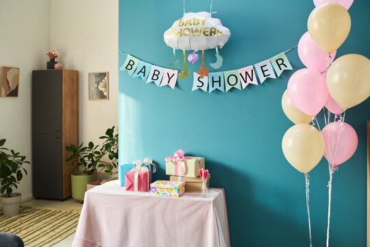 Decorated party table displaying wrapped gifts and pastel balloons against teal wall with baby shower banner, celebrating gender reveal event