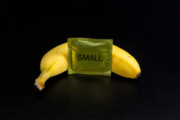 A small-sized condom next to a banana with a black background. Small peniss syndrome problem