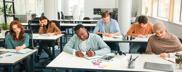 Modern Education Concept. Portrait of diverse group of students sitting at desks in classroom at university, taking test, entrance examination or writing notes in notebook, selective focus