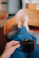 Relaxing at home with a cup of coffee, enjoying a peaceful morning A person relaxing with a mug of tea, feet up on a chair, casual indoor setting. A faceless woman sits on a chair sofa at cozy home.