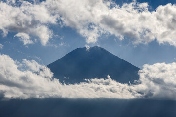 Mount Fuji surrounded by drifting clouds with beams of sunlight piercing through creating a...