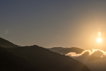 Golden sunlight rises over misty mountain ridges, casting long shadows and illuminating drifting clouds in a peaceful morning scene