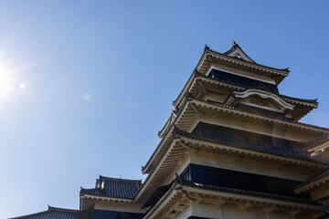 Matsumoto Castle tower in Nagano with layered tiled roofs and wooden panels is lit by sunlight from the left side with lens flare against clear sky