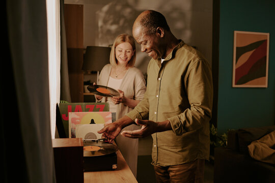 Senior Black man and senior Caucasian woman selecting vinyl records together, smiling and enjoying shared moment, standing near record player with jazz albums visible on table