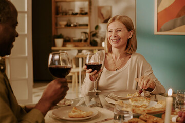Senior Caucasian woman smiling while sitting at dining table with senior Black man, both holding wine glasses and enjoying meal together in cozy home setting, expressing romance and companionship