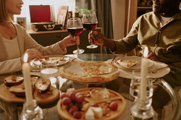 Senior Caucasian woman and senior Black man sitting at dining table clinking wine glasses, sharing romantic meal together, enjoying food and drink, smiling and making eye contact