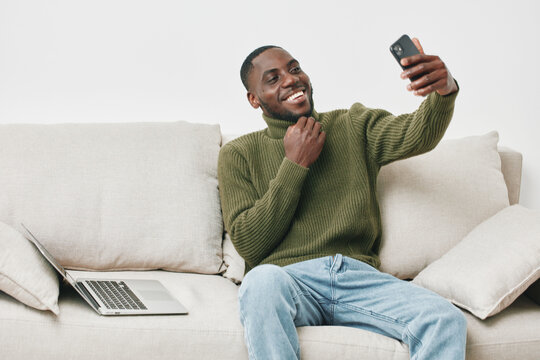 Smiling African American man in green sweater taking selfie on smartphone, relaxed on couch with laptop, modern home interior, cheerful and casual atmosphere