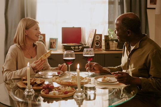 Senior Caucasian woman and senior Black man sitting at dining table sharing meal and wine, smiling at each other, enjoying romantic moment together in cozy home setting - Powered by Adobe