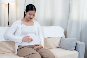 Asian pregnant woman sitting on sofa smiling touching belly wearing white sweater and brown pants in cozy living room expressing happiness excitement waiting for baby arrival soon