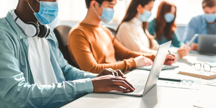 New Normal Reality Rules, People And Workspace Concept. Group of diverse international people wearing disposable surgical masks studying and working sitting at desk, preparing project, using laptop