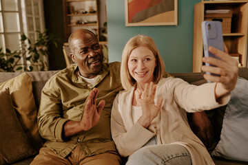 Senior Caucasian woman and senior Black man sitting on sofa smiling and waving while holding smartphone taking selfie together, showing romantic connection and joyful interaction
