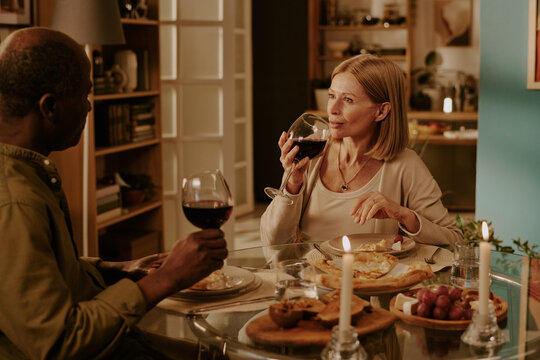 Senior Caucasian woman and senior Black man sitting at dining table sharing romantic dinner, holding wine glasses, engaging in conversation, enjoying intimate moment together