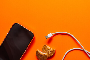 Flat lay of black smartphone, white charging cable and bitten cookies on bright orange surface, digital lifestyle and snack break pop art concept. High quality photo