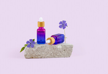 two bottles of essential oil with a pipette and periwinkle flowers on a stone podium on a purple...