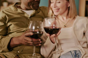 Senior Caucasian woman and senior Black man sitting close together holding wine glasses smiling and enjoying romantic moment, hands and faces partially visible, relaxed interaction
