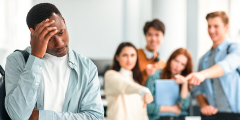 Bullying Concept. Rude diverse group of people mocking and making fun of sad african american guy, pointing fingers at him and laughing, standing in blurred background. Prank, abuse, public disgrace