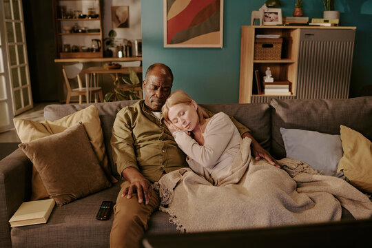 Senior Black man and senior Caucasian woman sitting on sofa, woman resting head on man's shoulder, couple embracing while relaxing together, remote control and book on couch - Powered by Adobe