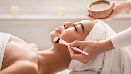 Beauty therapist applying face mask on young asian female skin at spa, professional beautician using brush for putting moisturising treatment to young korean lady face, side view, closeup