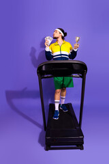 Young man in vibrant retro tracksuit holding cash and trophy while standing on treadmill against...