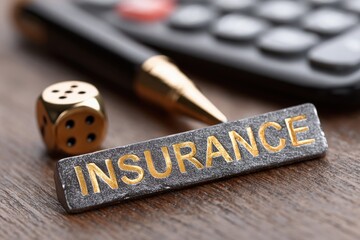 A calculator, gold pen, and dice are arranged next to an insurance plaque on wooden surface. The setting suggests a discussion on insurance options, risk management, and financial planning.