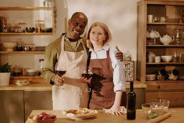 Senior Black man and senior Caucasian woman standing together in kitchen smiling and holding glasses of red wine, embracing each other while preparing meal on wooden counter