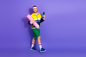 Young athletic man in retro-style activewear holding yoga mat and water bottle against a purple background