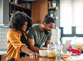 Happy young afro american couple having fun preparing food and looking for recipes online using a laptop in kitchen
