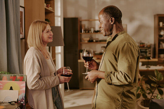Senior Caucasian woman and senior Black man standing together holding wine glasses, smiling and making eye contact, engaging in romantic conversation in cozy indoor setting