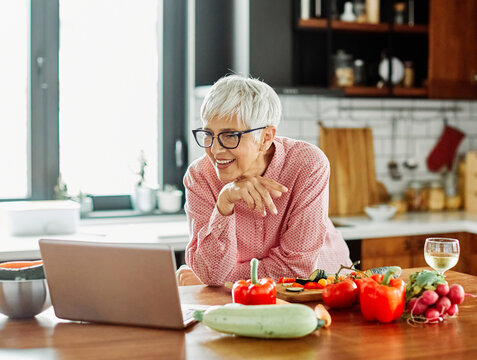 Portrait of happy senior mid aged mature woman prepering meal with fresh vegatebles and following internet instructions for a recipe on a laptop computer  or looking at video  or website app and drink
