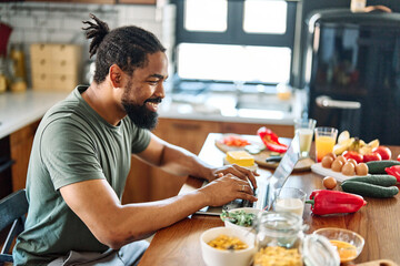 Happy young afro american man having fun preparing food and looking for recipes online using a laptop in kitchen, or a young businessman working from home office