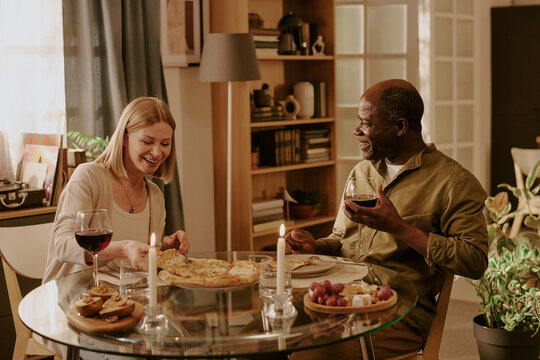 Senior Black man and senior Caucasian woman sitting at round glass table sharing romantic dinner with wine and pizza, smiling and looking at each other in cozy home setting