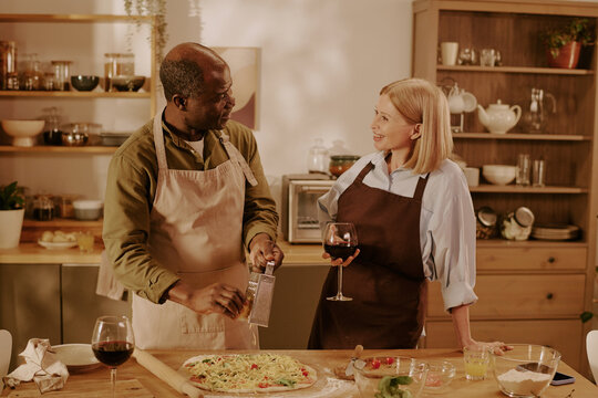 Senior Black man and senior Caucasian woman preparing meal together in kitchen, smiling and making eye contact, man grating cheese onto pizza while woman holding glass of red wine - Powered by Adobe