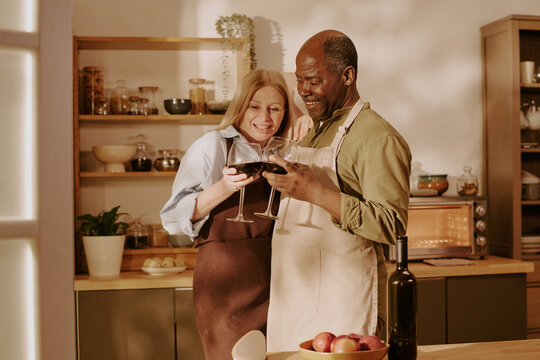 Senior Caucasian woman and senior Black man standing together in kitchen smiling and holding wine glasses, embracing and enjoying romantic moment, kitchen shelves in background
