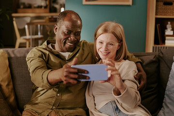 Senior Caucasian woman and senior Black man sitting close together on sofa smiling and taking selfie with smartphone, showing affection and enjoying romantic moment at home