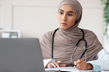 Muslim female doctor having professional online training on laptop, taking notes while watching...