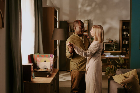Senior Black man and senior Caucasian woman dancing together in living room, holding hands and looking at each other, expressing romance and connection in home setting