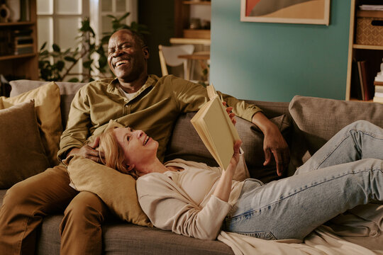 Senior Caucasian woman lying on sofa holding book smiling up at senior Black man sitting beside her laughing, couple relaxing together in living room showing romantic connection