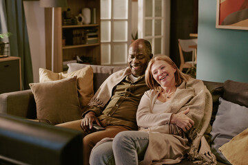 Senior Caucasian woman and senior Black man sitting close together on sofa smiling and watching television, woman leaning affectionately on man, couple sharing romantic moment at home